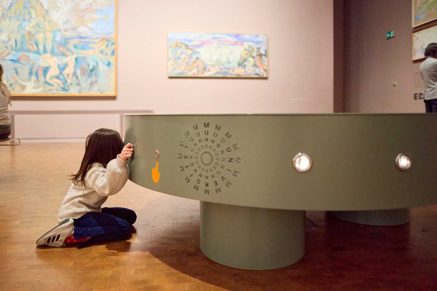 En ung jente sitter på kne og ser inn gjennom et kikkhull i en installasjon i ustillingen Monumental.