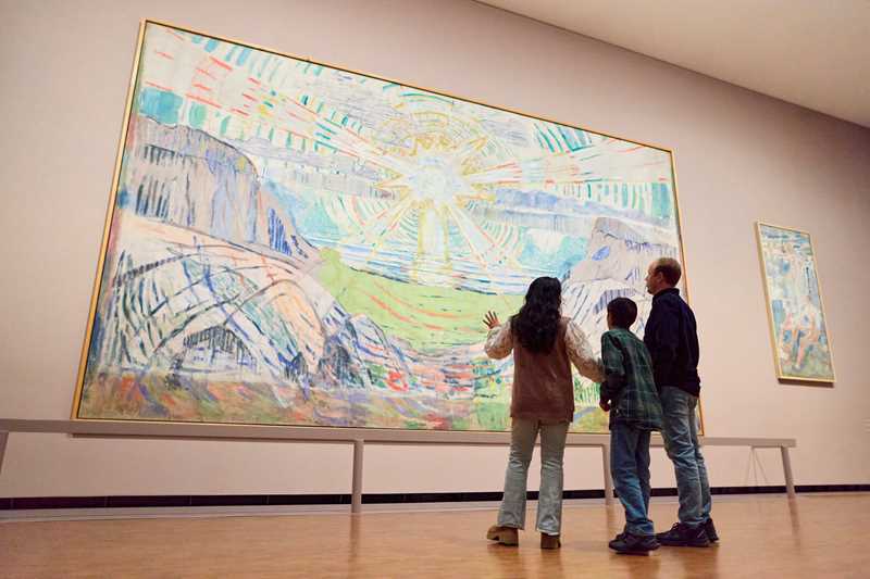 A mother, son and father stand looking at The Sun by Edvard Munch. They are facing away from the camera. Photograph.