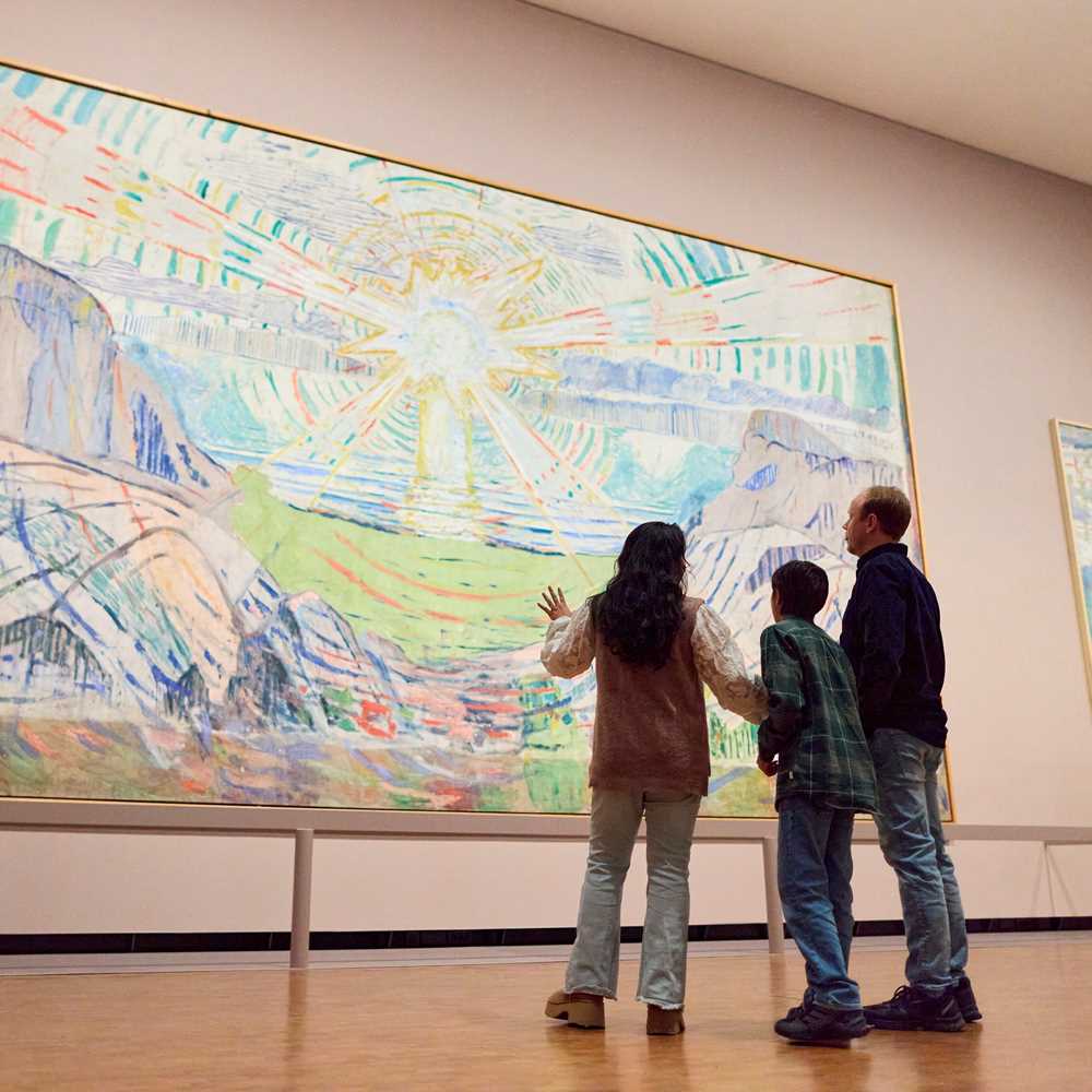 A mother, son and father stand looking at The Sun by Edvard Munch. They are facing away from the camera. Photograph.