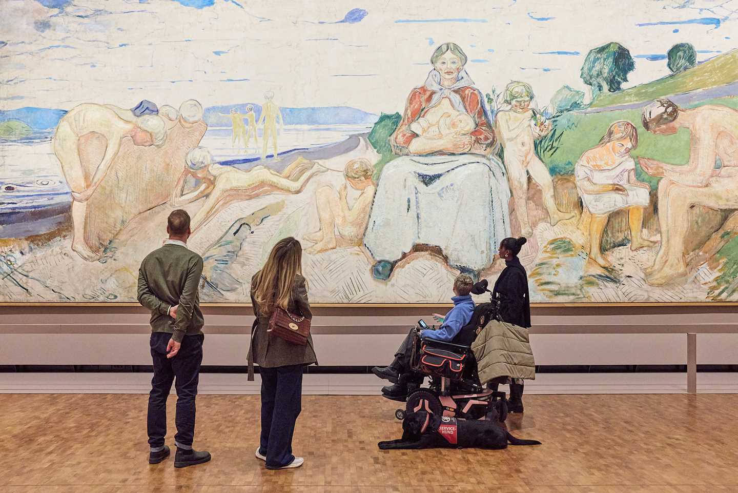 Fire personer står med ryggen mot kamera og betrakter maleriet Alma Mater av Edvard Munch. Bildet er 50 kvadratmeter stort. Fotografi