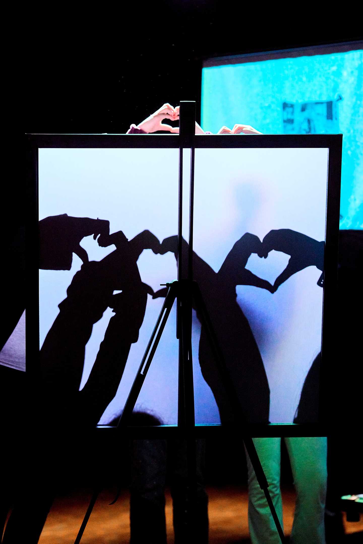 Three teenagers stand behind an illuminated screen, making heart-shaped shadow figures with their hands. Photograph.