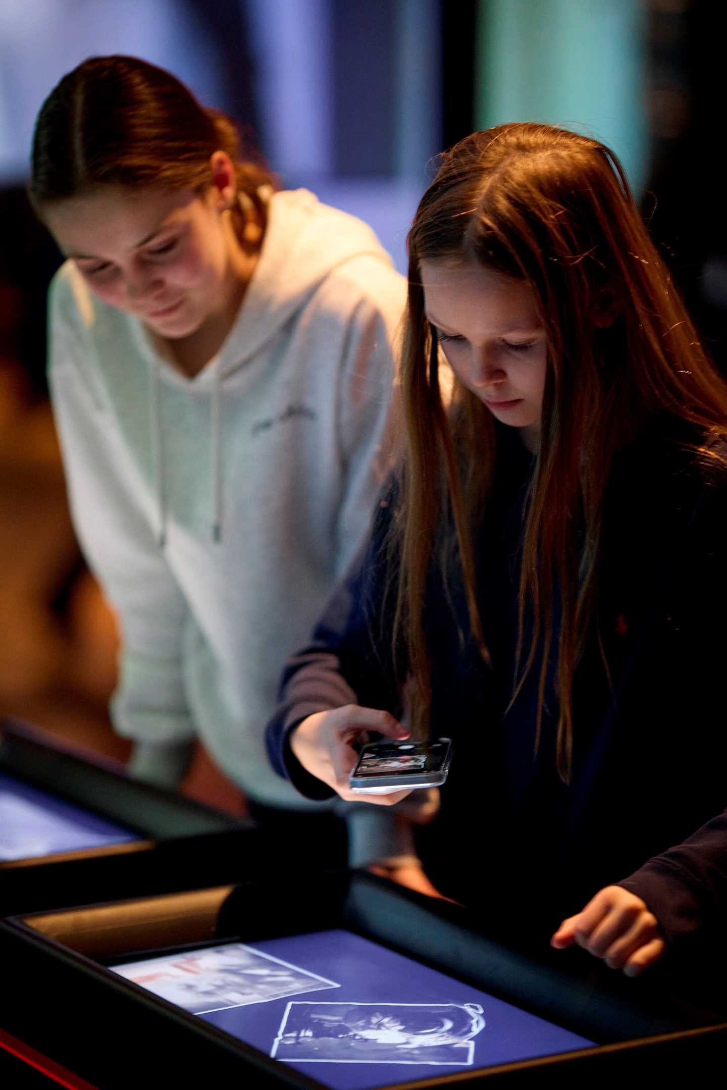 Two girls look down at a digital screen displaying an image. One of the girls takes a photo of the image with her phone. Photograph