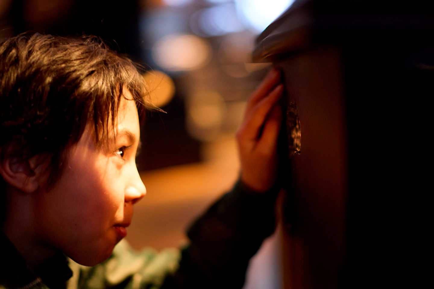 A boy crouches and looks through a glowing mouse hole in the wall. Photograph.