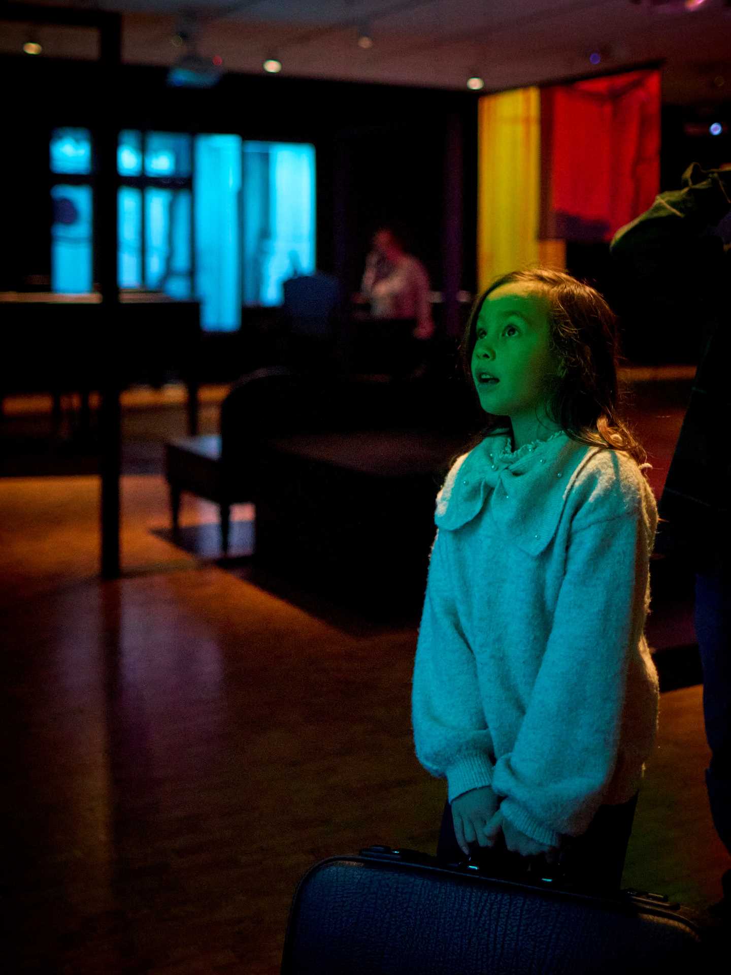 A young girl stands holding a suitcase. Her face is lit with a green glow, and she looks mesmerised by something outside the frame. Photograph.