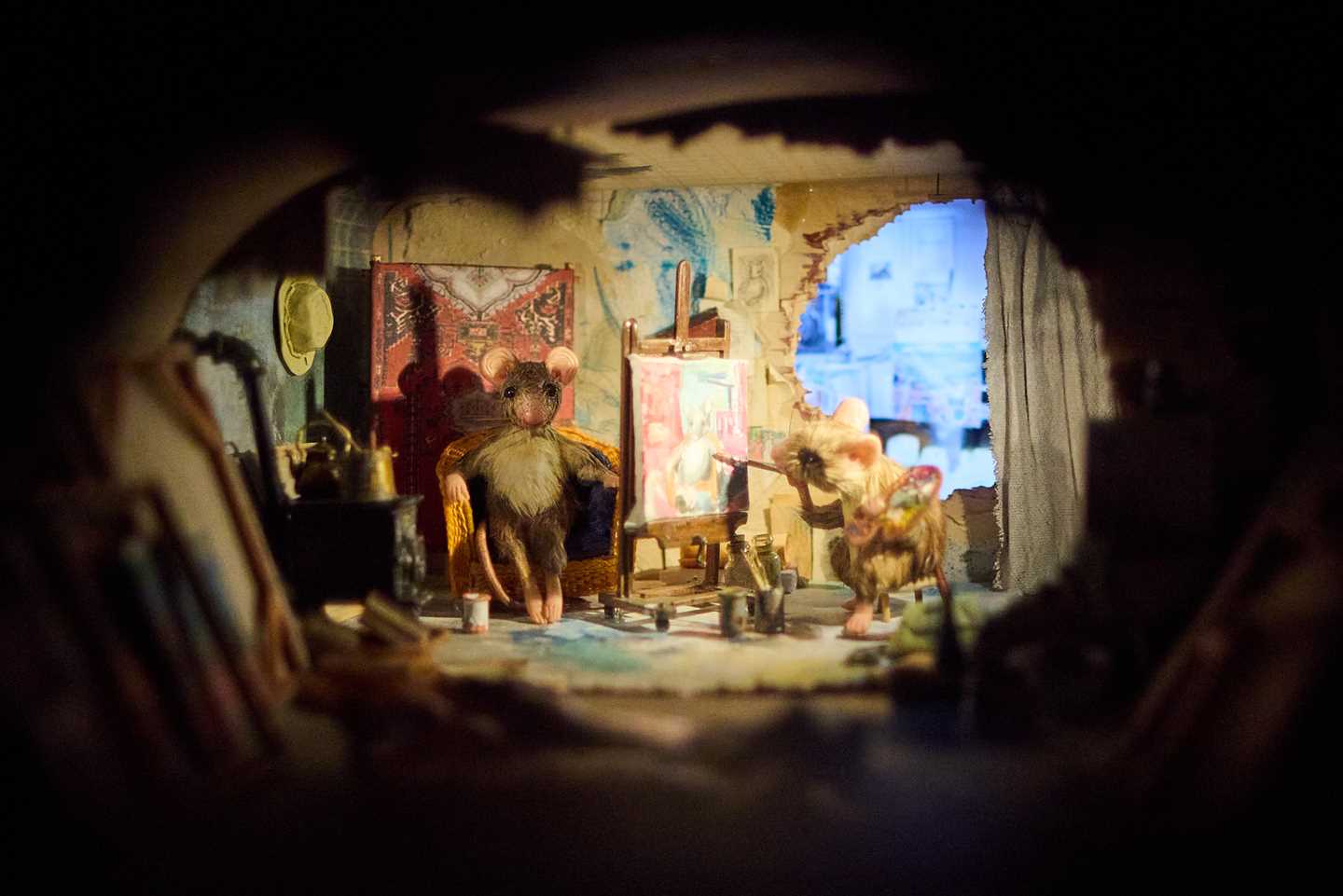 View into a mouse hole revealing a cosy room where one mouse paints at an easel while another poses in a wicker chair. Photograph.
