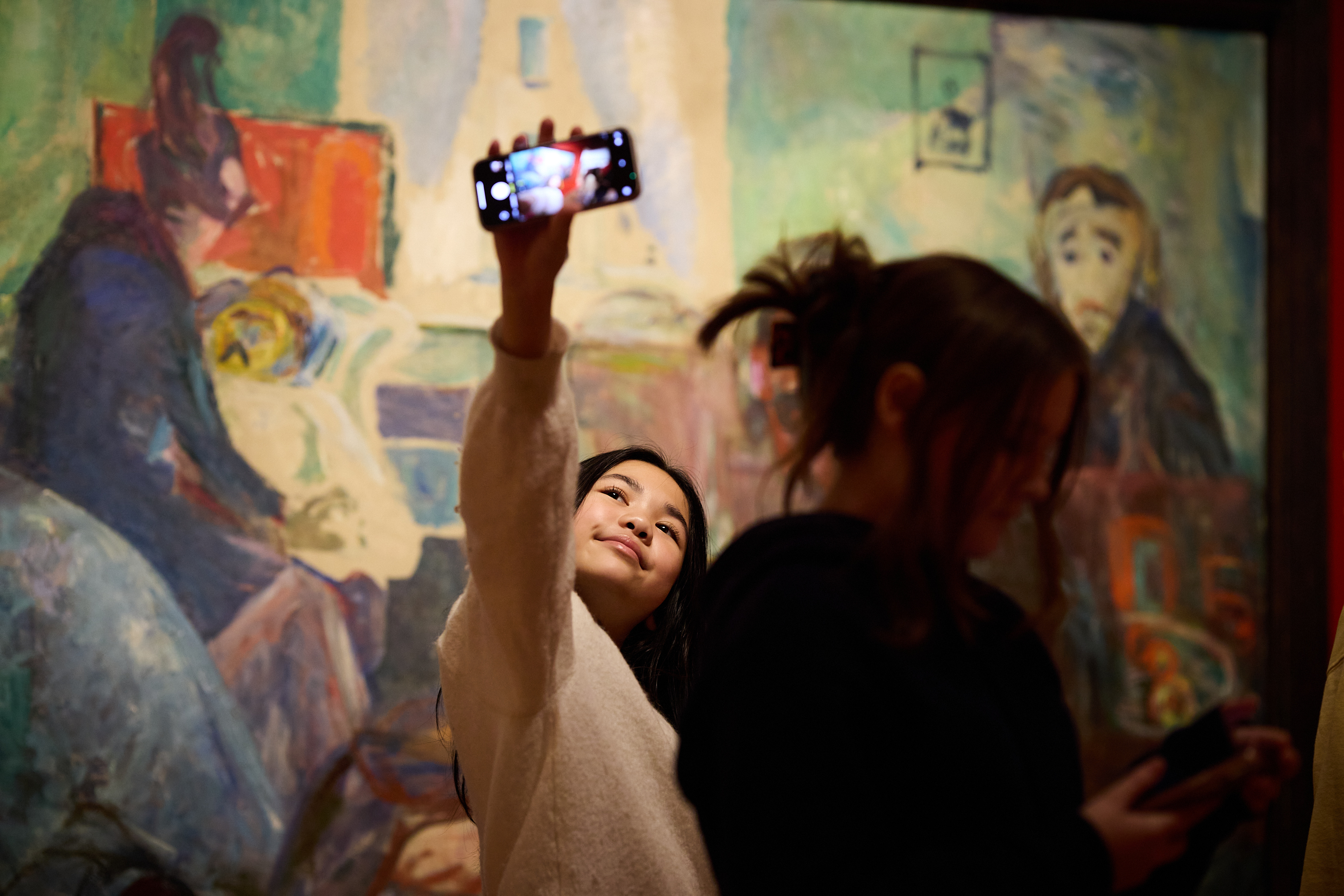 En ung jente tar et bilde av seg selv med mobilen sin foran et maleri av Edvard Munch i utstillingen Uendelig. Fotografi