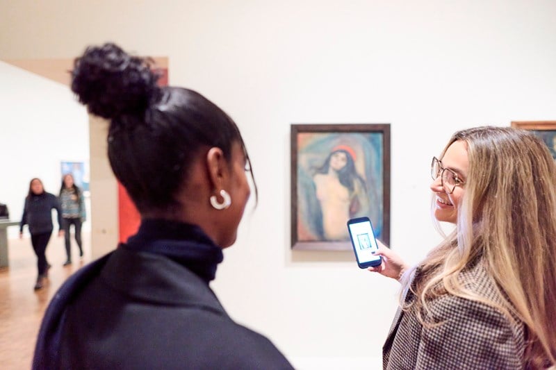 To kvinner snakker sammen mens de ser på maleriet Madonna av Edvard Munch. Den ene kvinnen tar et bilde av maleriet med mobilen sin. Fotografi