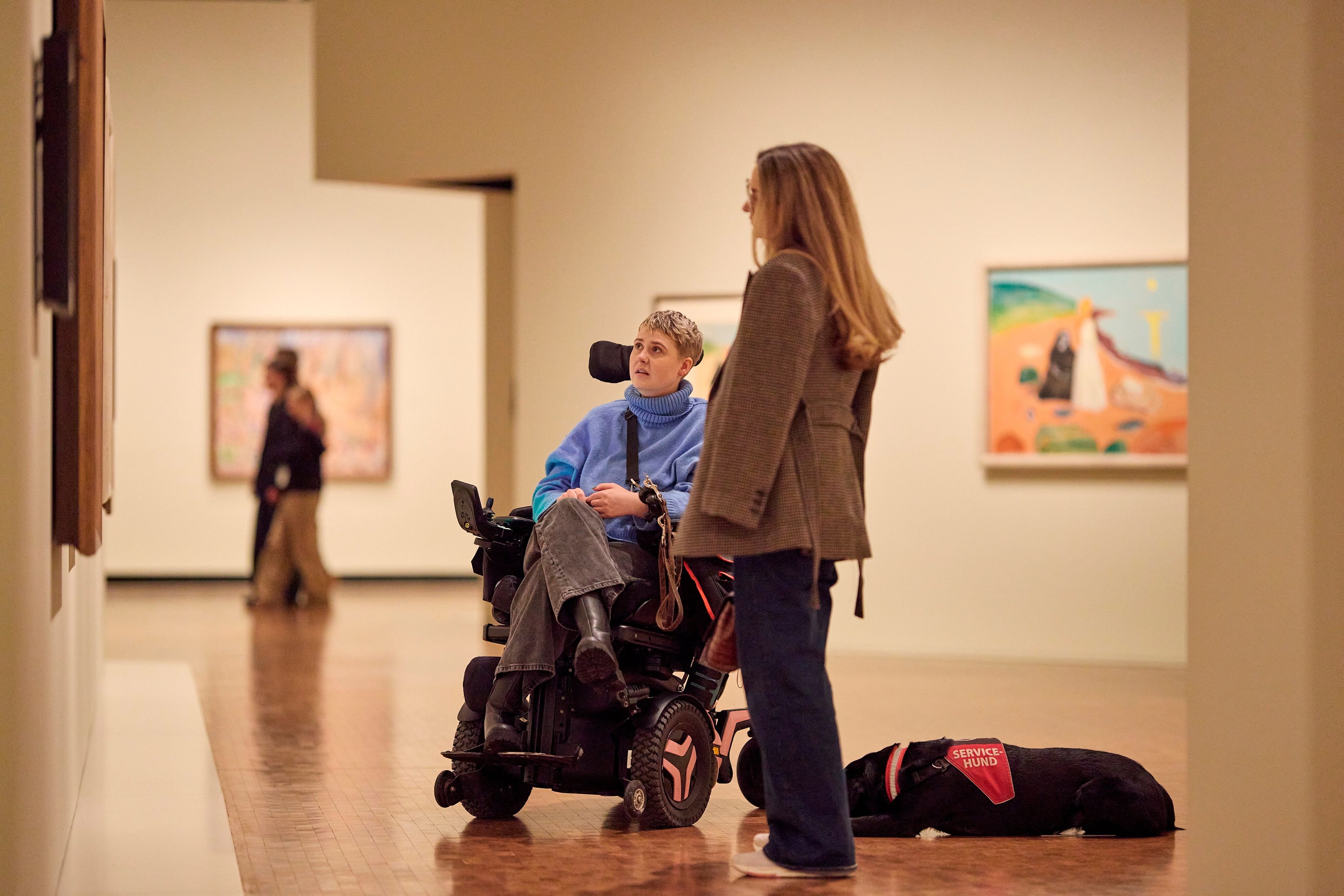 Two friends admiring a work by Edvard Munch in the Infinite exhibition. One woman is in a wheelchair, and a service dog lies on the floor behind them. Photograph.