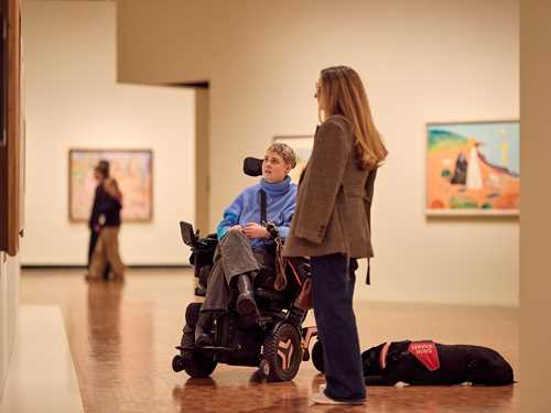 Two friends admiring a work by Edvard Munch in the Infinite exhibition. One woman is in a wheelchair, and a service dog lies on the floor behind them. Photograph.