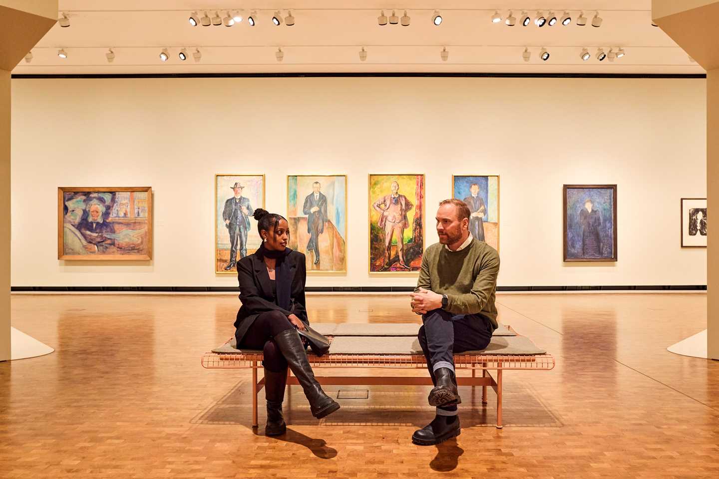 En kvinne og en mann sitter på en benk inne i en utstillingssal og snakker sammen. På veggen bak dem henger flere portrettmalerier av Edvard Munch. Fotografi