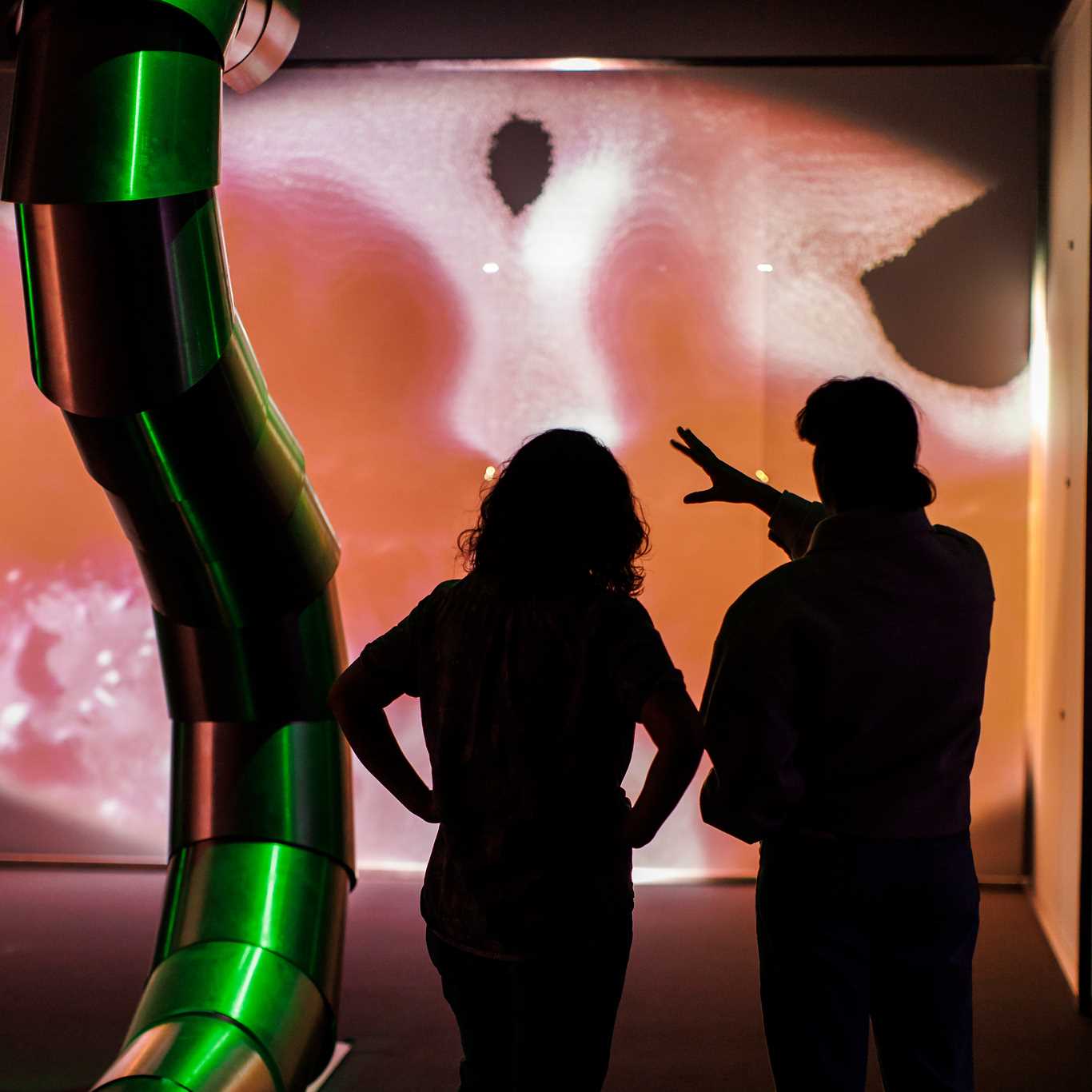 Two people stand in an exhibition hall, viewing artwork by Sandra Mujinga. They are seen from behind. Photograph.