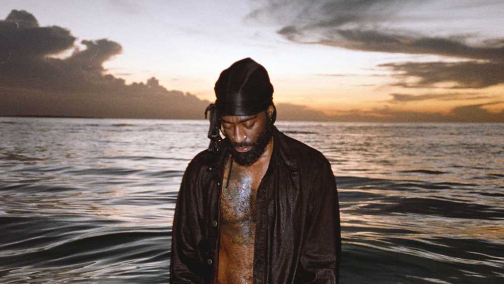 Arif stands in the sea with water up to his thighs, looking down into the water. He is wearing an open black shirt, and there is a sunset in the background. Photograph.