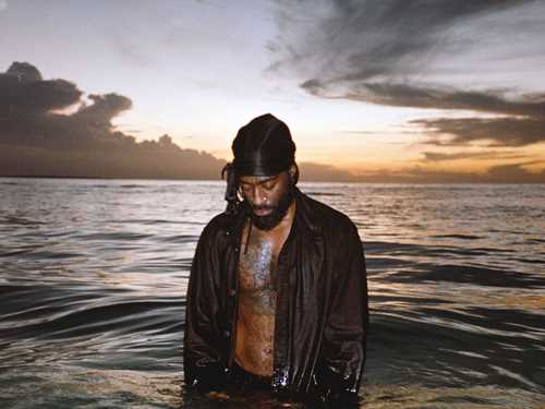 Arif stands in the sea with water up to his thighs, looking down into the water. He is wearing an open black shirt, and there is a sunset in the background. Photograph.