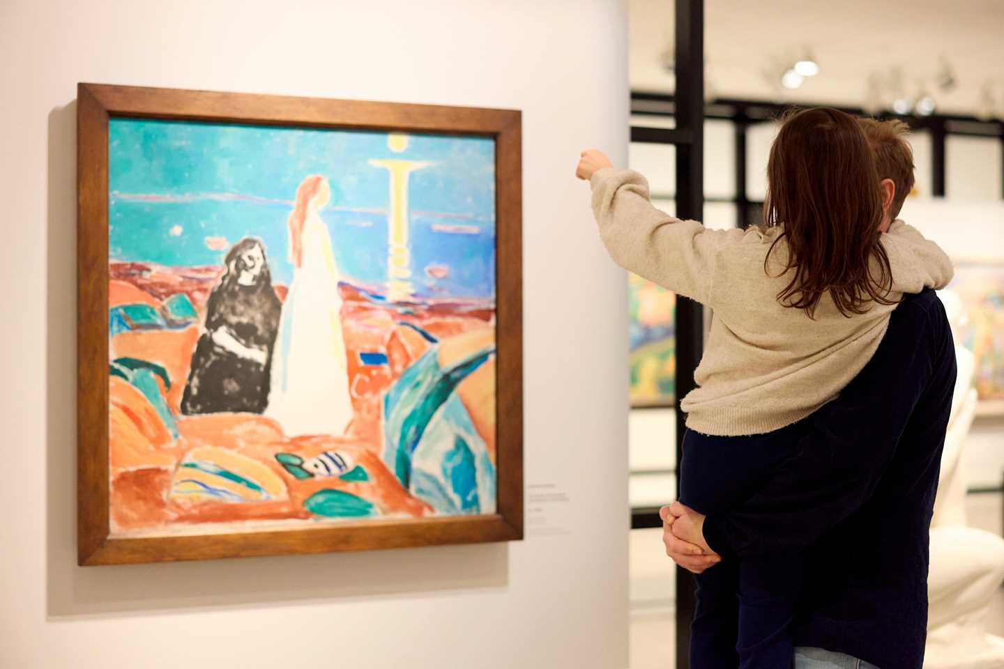 A father holds his daughter in his arms, both with their backs to the camera. They are looking at a painting by Edvard Munch. Photograph.