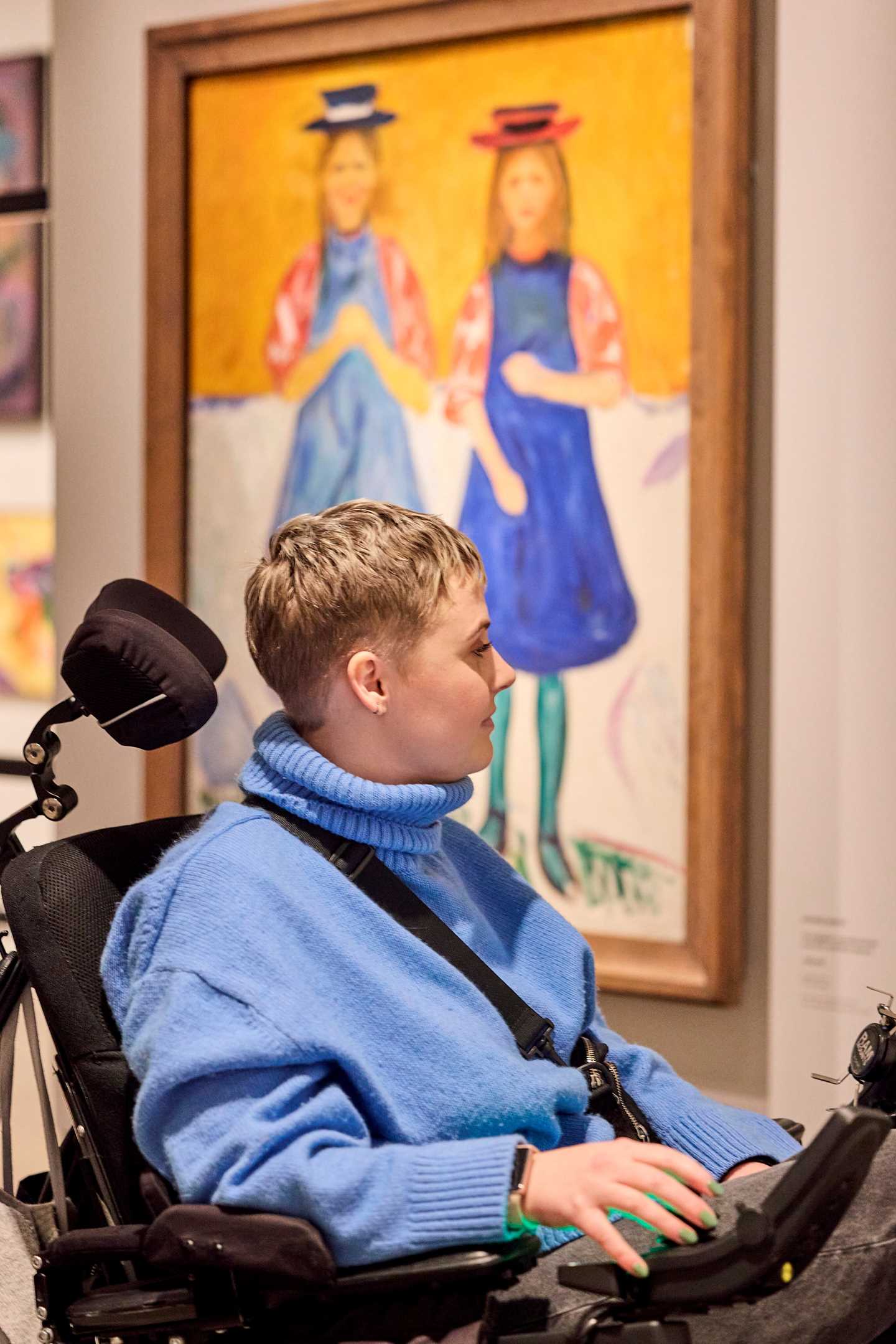 A woman in a wheelchair, seen from the side, looks at an artwork. Behind her is the painting Two Girls with Blue Aprons. Photograph.