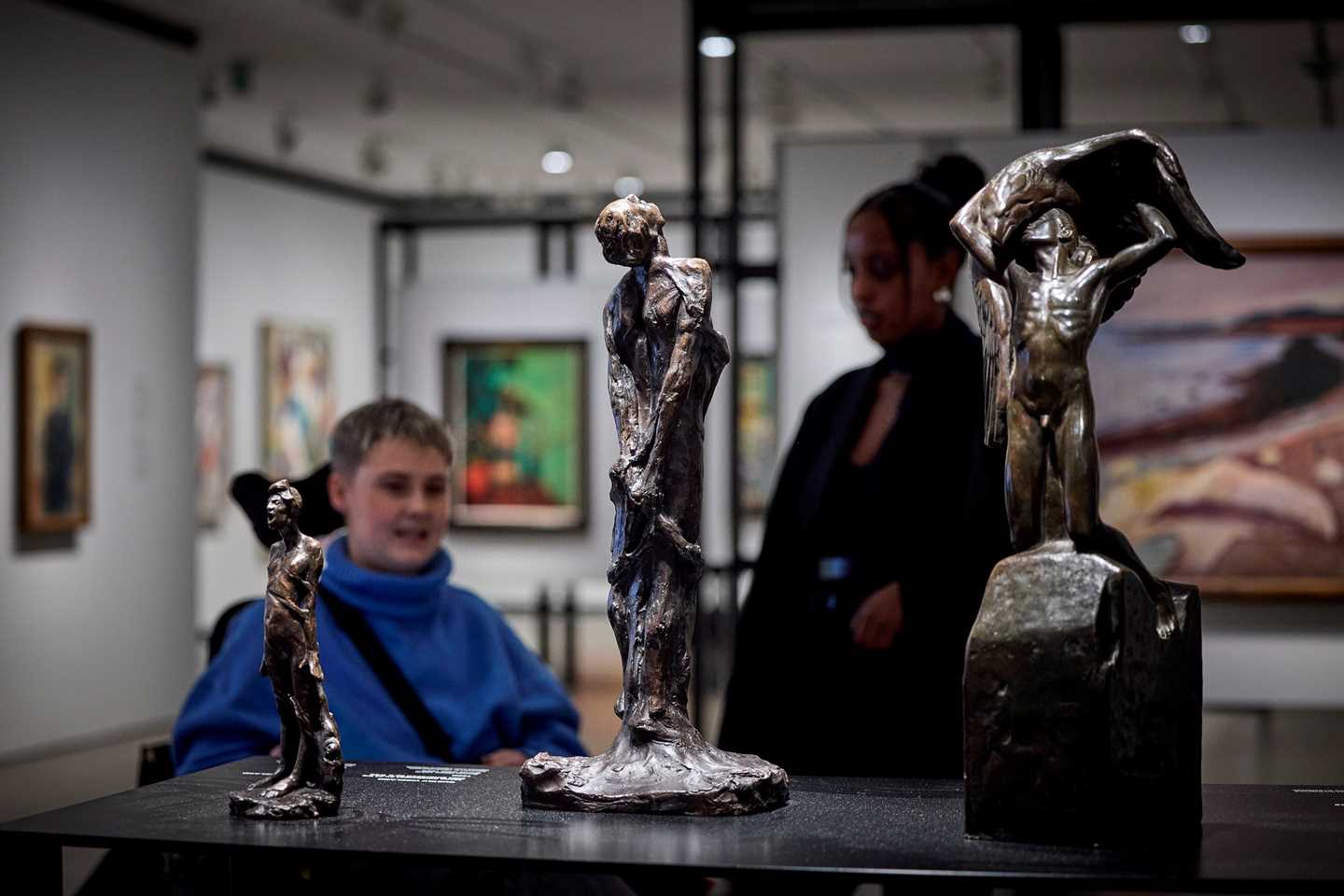 Three bronze sculptures of people in different poses. In the background, two people are talking, with the rest of the exhibition room visible. Photograph.
