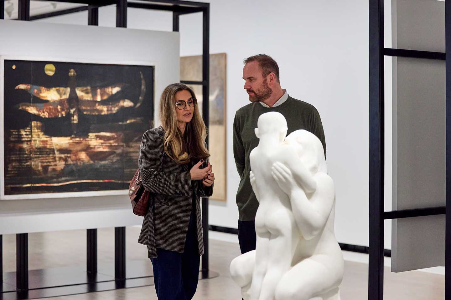 A woman and a man stand in an exhibition room having a conversation. In front of them is a white sculpture of a woman and a child. Behind them, an artwork hangs on the wall. Photograph.