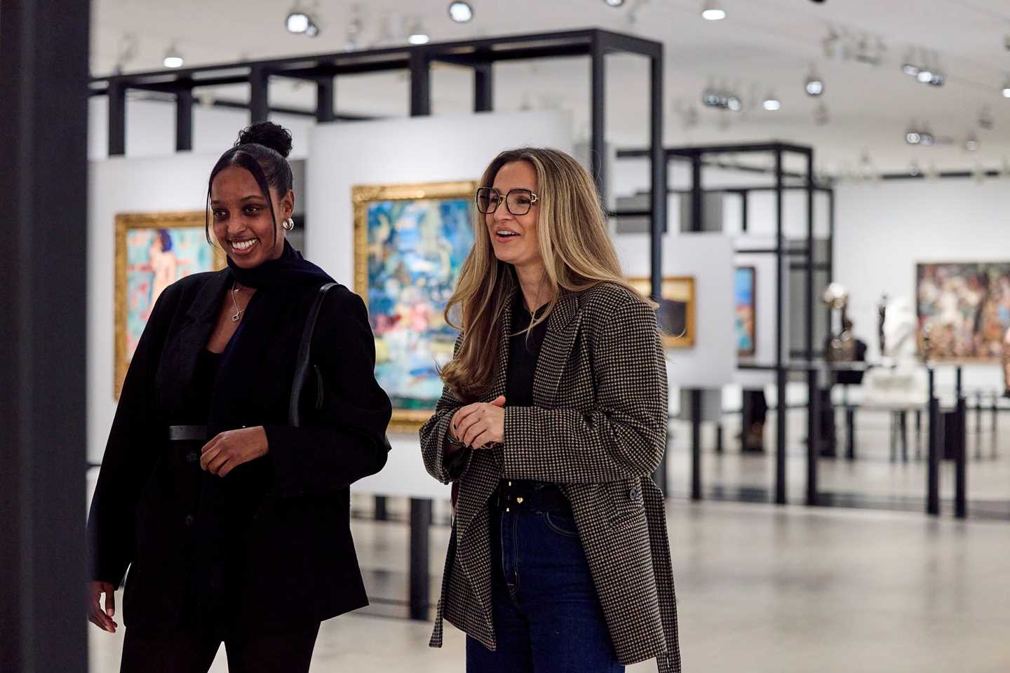 Two women stand looking at an artwork. Behind them is an exhibition room with black structures displaying artworks. Photograph.