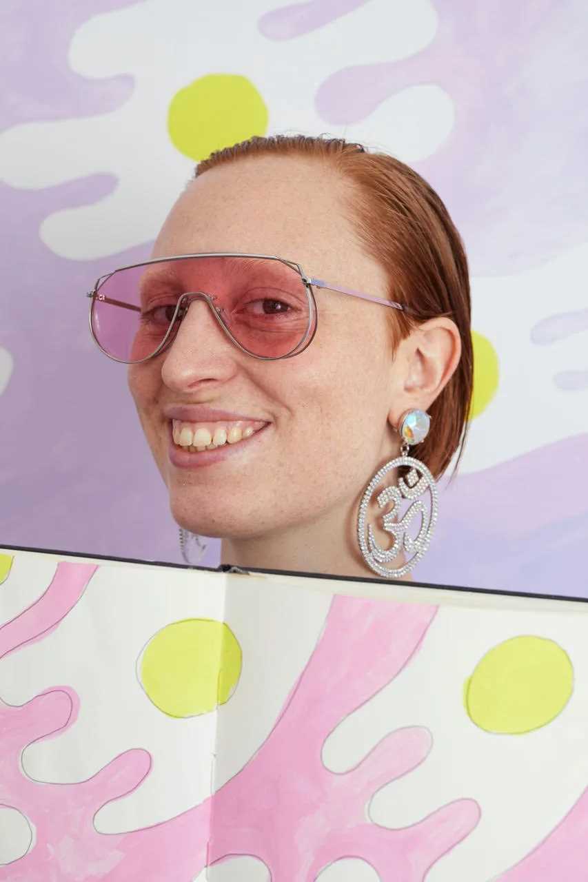 Constance Tenvik med rosa solbriller smilende med en skissebok. Fotografi.