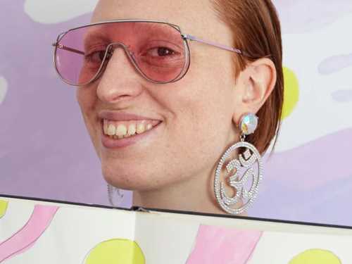 Constance Tenvik wearing pink sunglasses, smiling with a sketchbook. Photograph.
