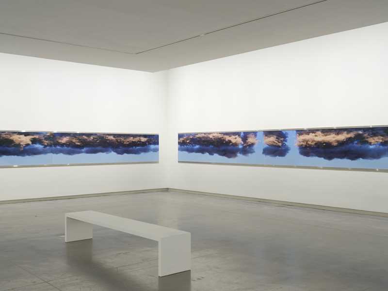 A white exhibition room with a white bench in the centre. Along the walls hang continuous narrow strips showing blue sky and clouds. Photograph.