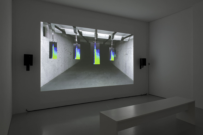 A small white room with a projection on the wall showing sound frequencies. On each side of the projection there is a black speaker, and on the floor in front of the projection there is a white bench. Photography