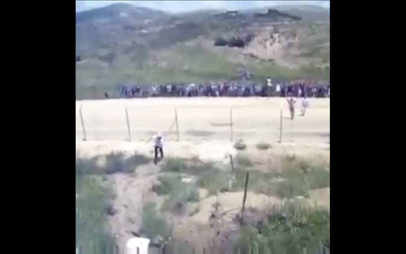 A heavily pixelated photograph showing mountain formations in the background, a sandy plain with a crowd of people at one end, and a wire fence at the other. One person stands on the same side of the fence as the camera, while the rest are on the other side. Photograph.