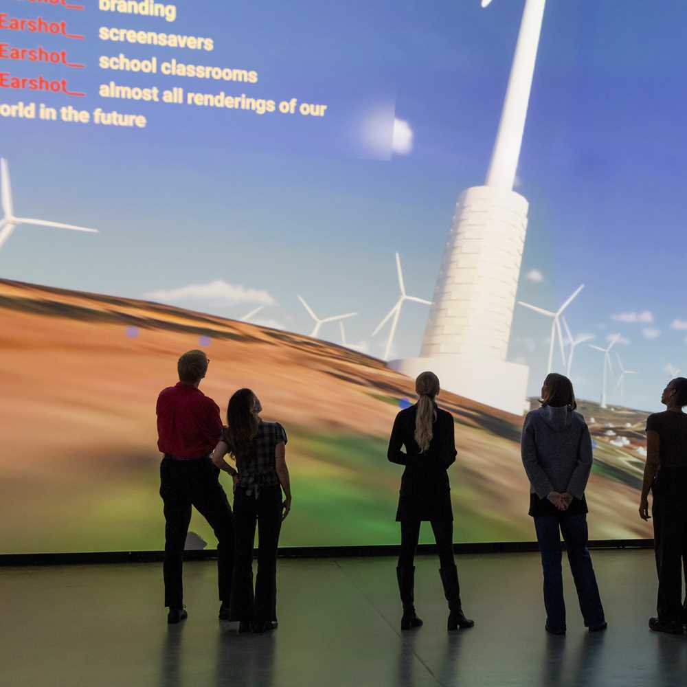 En gruppe mennesker står inne i et mørkt rom foran en vegg som er dekket av en projeksjon som viser en dataanimasjon av vindmøller på Golanhøydene. Fotografi