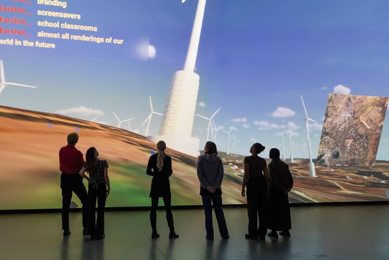 En gruppe mennesker står inne i et mørkt rom foran en vegg som er dekket av en projeksjon som viser en dataanimasjon av vindmøller på Golanhøydene. Fotografi