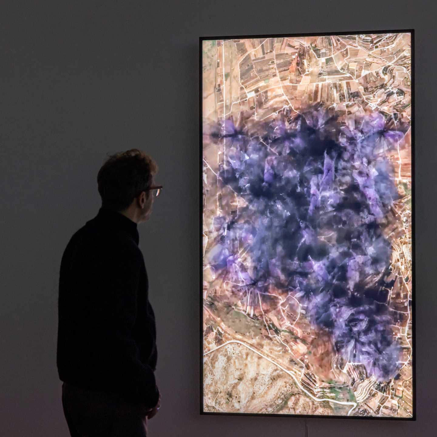Kunstneren Lawrence Abu Hamdan står i et mørkt rom og ser på en lodrett skjerm som viser en visualisering av støyen 31 vindturbiner vil skape på Golanhøydene. På skjermen ser du at støyen sprer seg utover et stort område som lilla og svart gnistrende røyk. Fotografi