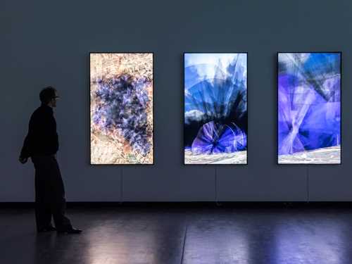 Kunstneren Lawrence Abu Hamdan står inne i et mørkt rom foran tre vertikale skjermer som henger på veggen og viser visualiseringer av støyen 30 vindturbiner vil skape på Golanhøydene. Støyen brer seg ut som sort og lilla røyk. Fotografi