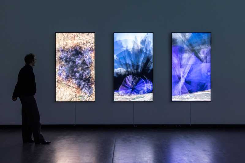 The artist Lawrence Abu Hamdan stands in a dark room in front of three vertical screens mounted on the wall, showing visualisations of the noise that 30 wind turbines would generate on the Golan Heights. The noise spreads out like black and purple smoke. Photograph