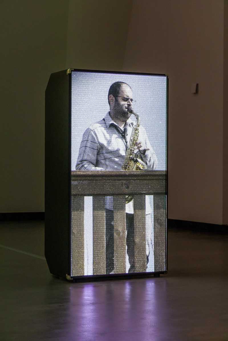 A projection of the saxophonist Amr Mdah, playing while standing on a balcony, is displayed on a loudspeaker. Photograph
