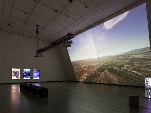 Et mørkt utstillingsrom som blir lyst opp av en projeksjon som dekker hele den ene veggen i rommet. Projeksjonen viser en dataanimasjon av vindturbiner på Golanhøydene. Midt i rommet står en svart benk og på veggen til venstre i bildet henger tre vertikale skjermer. Helt til høyre i bildet ser man en høyttaler med en projeksjon av saksofonisten Amr Mdah som spiller fra en balkong. Fotografi