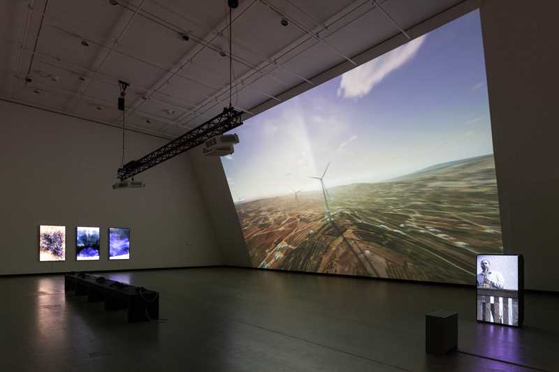 A dark exhibition room illuminated by a projection covering an entire wall. The projection shows a computer animation of wind turbines on the Golan Heights. In the middle of the room stands a black bench, and on the wall to the left of the image hang three vertical screens. On the far right of the image, a loudspeaker displays a projection of the saxophonist Amr Mdah playing from a balcony. Photograph