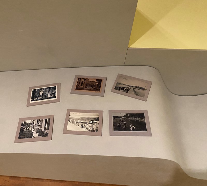 A photograph of six objects placed on moulded furniture within the exhibition space. The objects are reproductions of vintage postcards on small handheld boards. The postcards show different spas, seaside resorts and other destinations in Europe connected to health and wellbeing.