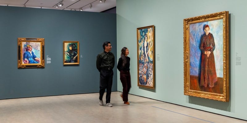 To mennesker går gjennom en utstilling og ser på malerier av Ludvig Karsten mens de smiler. Fotografi