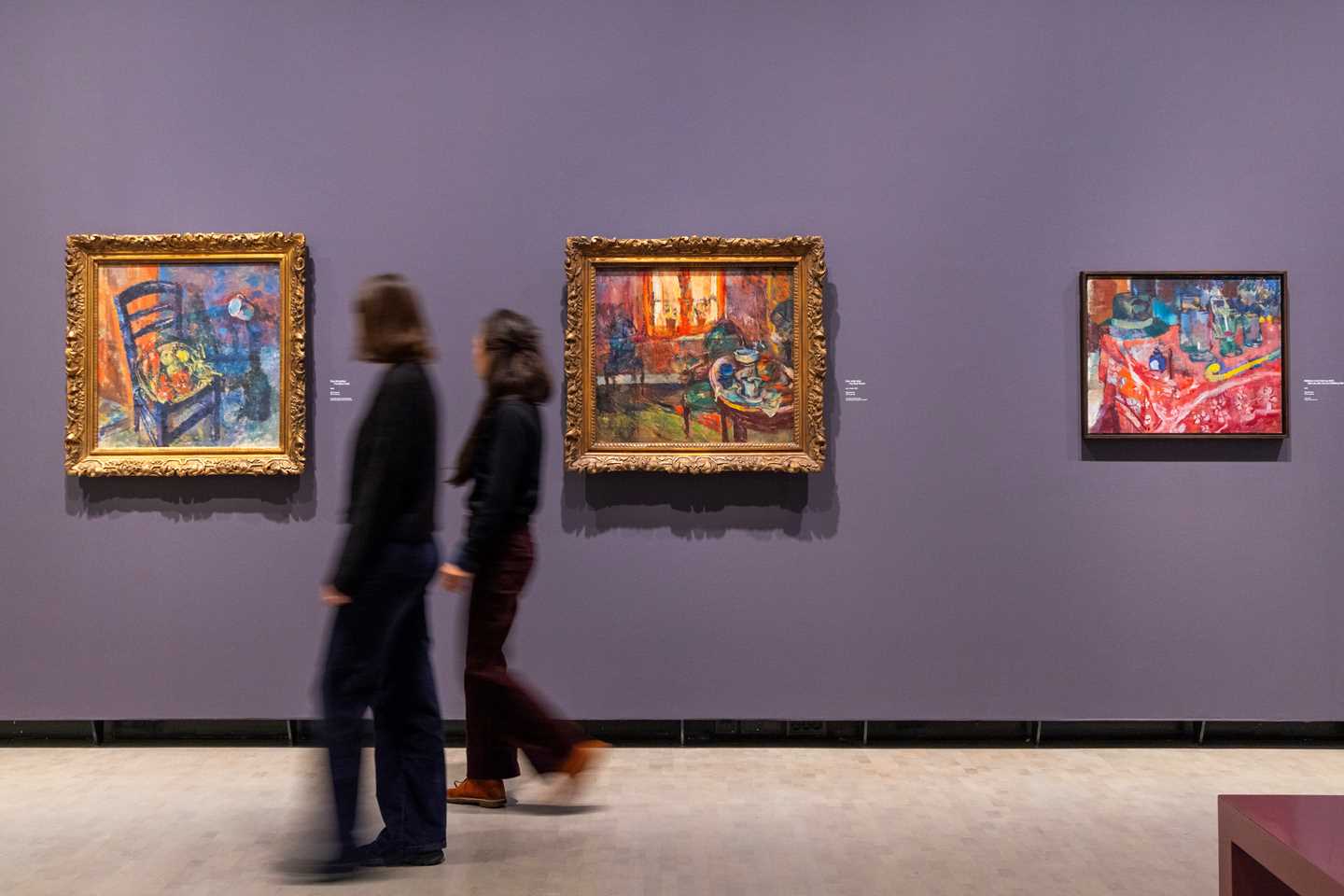 To kvinner går langs en lilla vegg med tre malerier av Ludvig Karsten. Fotografi.
