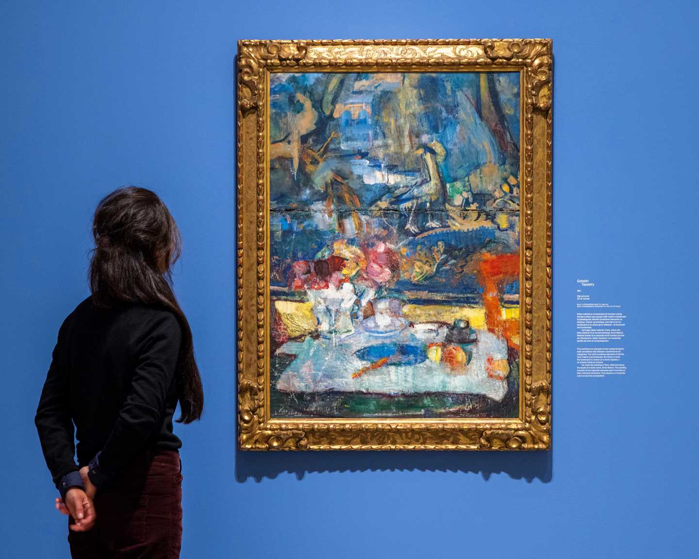 En kvinne ser på verket Gobelin av Ludvig Karsten. Maleriet henger mot en blå vegg og har en kraftig gullramme. Fotografi