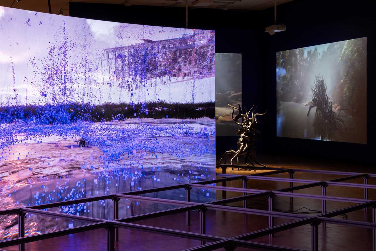 I forkant står en buet skjerm som viser et dystopisk landskap. I bakgrunnen ser vi skulpturer og tilhørende videoverk av Sahej Rahal. Fotografi