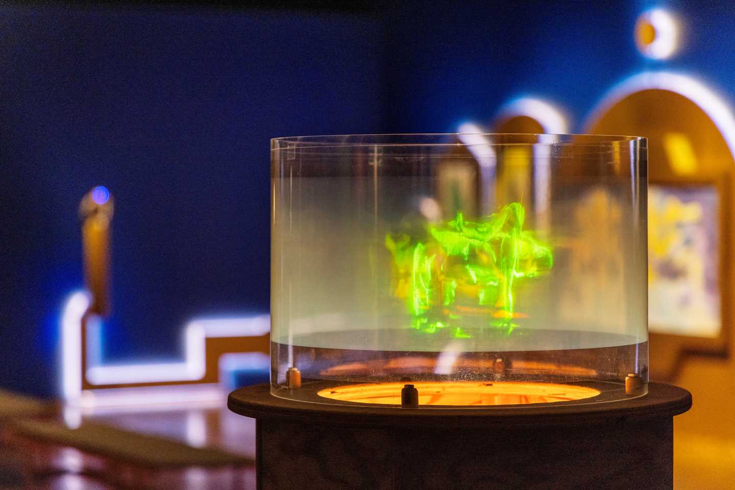 Et hologram som viser dansende, grønne mennesker er i fokus i bildet. Bak ser vi et blått utstillingsrom og en annen kunstinstallasjon. Fotografi