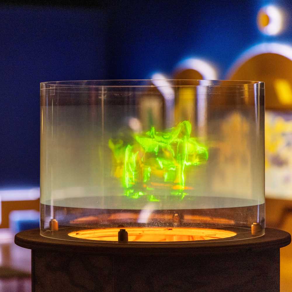 Et hologram som viser dansende, grønne mennesker er i fokus i bildet. Bak ser vi et blått utstillingsrom og en annen kunstinstallasjon. Fotografi