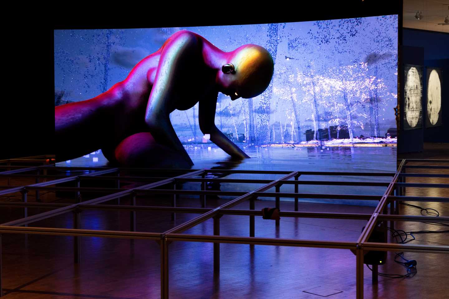 En stor buet skjerm viser en dataanimert video og på gulvet rundt skjermen står en metallkonstruksjon, bestående av et rutenett med påmonternte høyttalere. Verket er skapt av kunstnerkollektivet Infopsin. Fotografi 