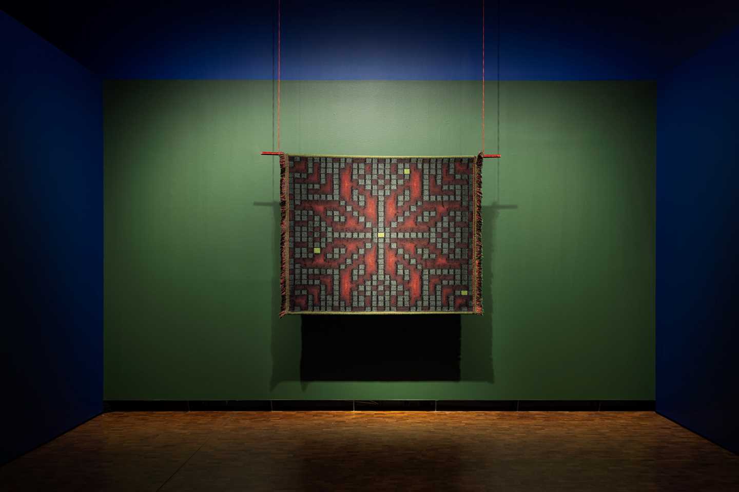 En kvadratisk vev henger mot en grønn bakgrunn. Mønsteret i veven minner om selburosen, men er satt sammen av små bokser som estetisk minner om klosser fra tetris eller lignende dataspill. Fotografi