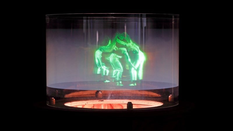 Et hologram viser mennesker som danser sammen i en ring. Menneskene er grønne og lyser opp bildet som ellers er helt mørkt. Fotografi