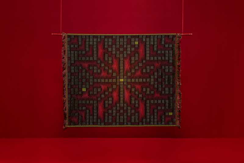 En kvadratisk vev henger mot en rød bakgrunn. Mønsteret i veven minner om selburosen, men er satt sammen av små bokser som estetisk minner om klosser fra tetris eller lignende dataspill. Fotografi