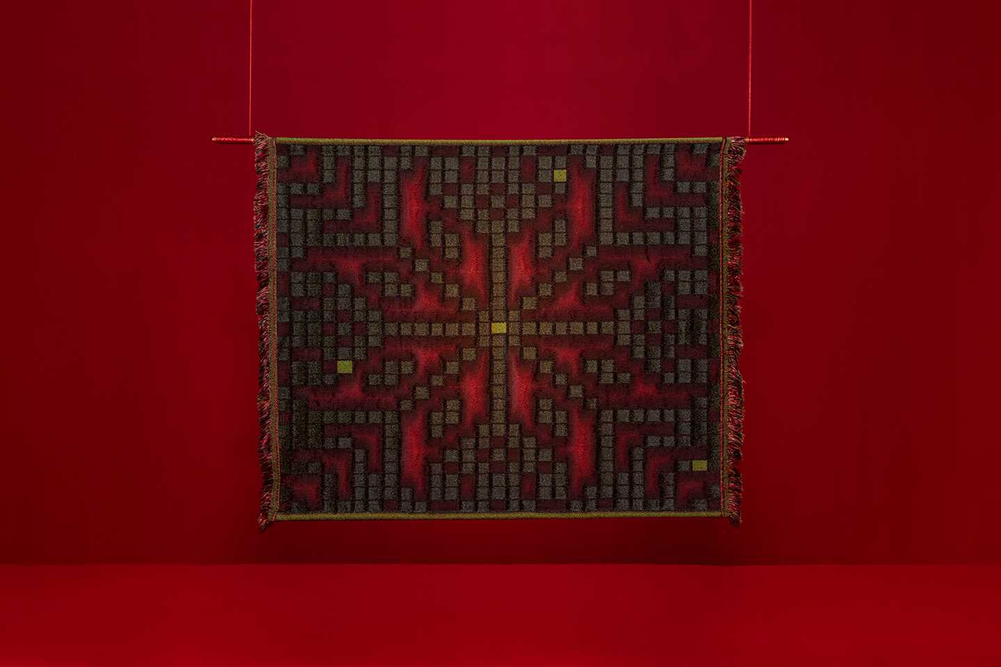 En kvadratisk vev henger mot en rød bakgrunn. Mønsteret i veven minner om selburosen, men er satt sammen av små bokser som estetisk minner om klosser fra tetris eller lignende dataspill. Fotografi