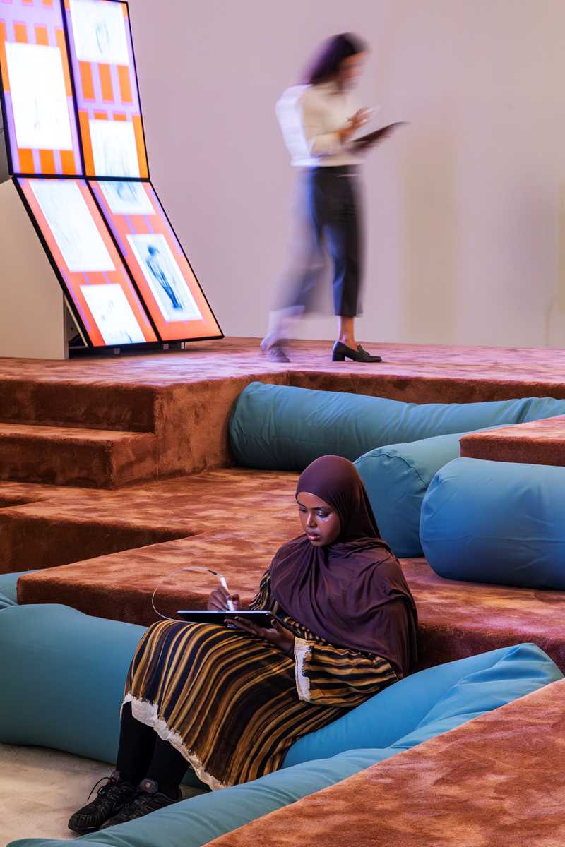 Woman seated on a beanbag drawing on an iPad, with another woman standing in the background, also drawing on an iPad. Photograph.