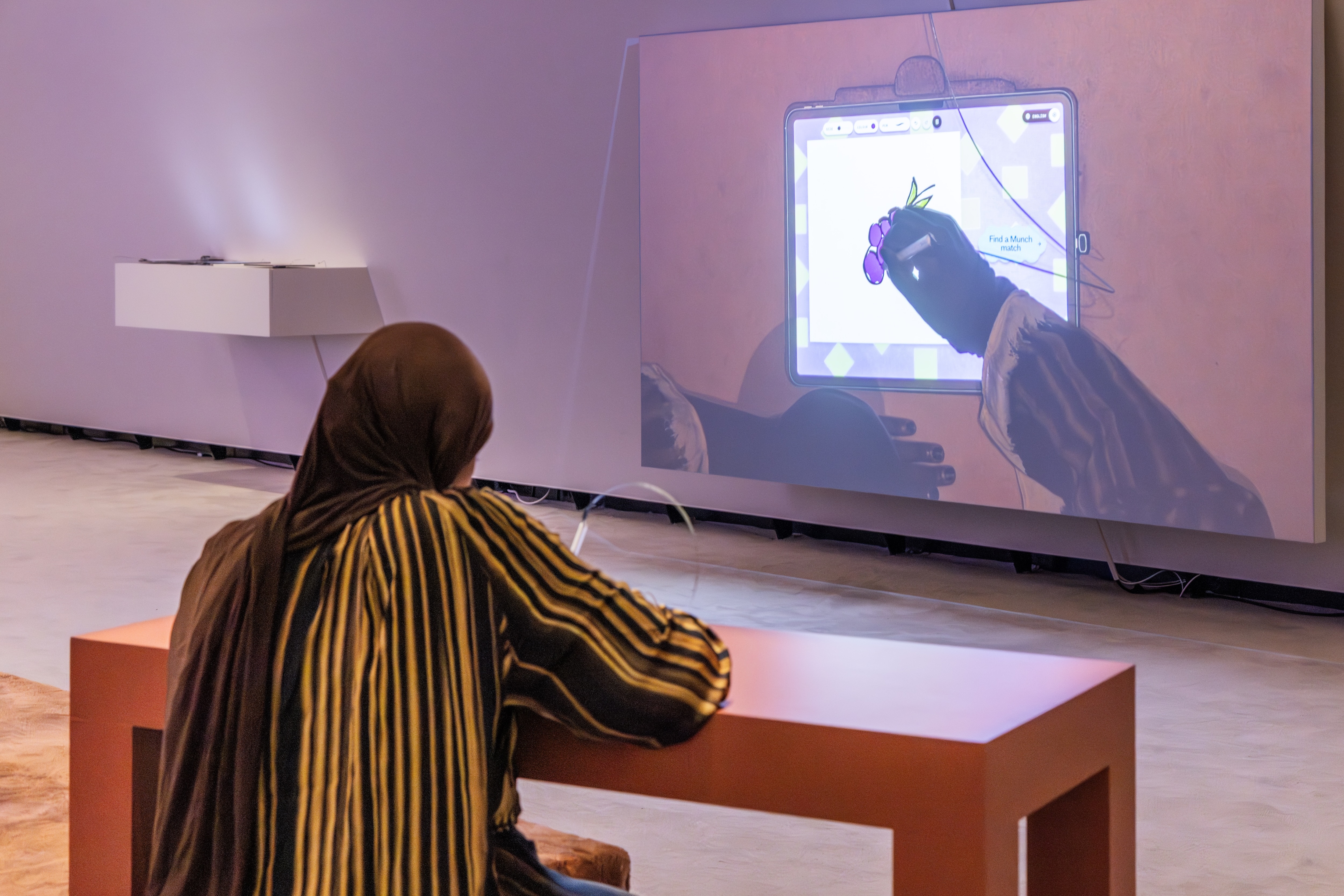 Woman seated at a desk drawing on a screen. An overhead camera projects her hands and drawing onto a wall so others in the gallery can watch in real time. Photograph.