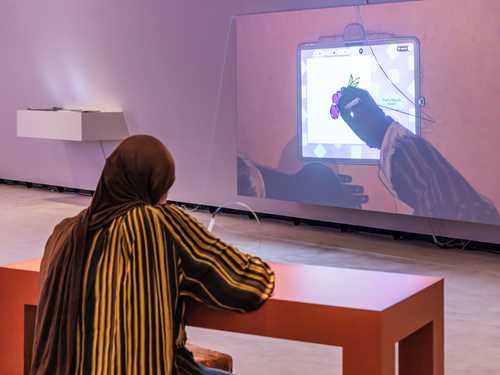 Woman seated at a desk drawing on a screen. An overhead camera projects her hands and drawing onto a wall so others in the gallery can watch in real time. Photograph.